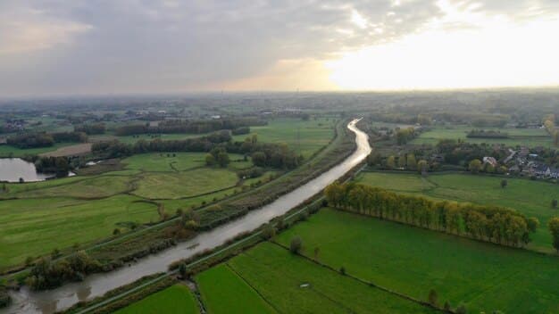 Aerial view of the Padma Bridge surrounded by green fields and rural settlements. The river is wide and calm, with boats sailing underneath the bridge. The sky is slightly cloudy.